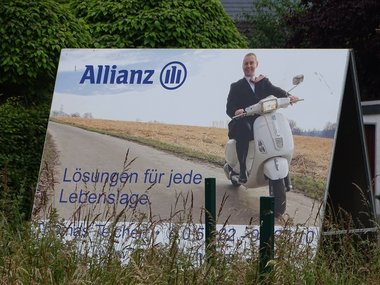 Ein Werbeplakat der Allianz mit einem Mann auf einem Roller auf einem Feldweg, der Aufschrift: Lösungen für jede Lebenslage, steht im Grünen von Gräsern und Büschen umgeben.