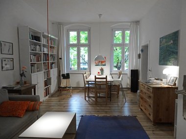 Sehr gemütliches Wohn- und Esszimmer in einer Berliner Altbauwohnung mit Holzparkett und warmem Licht. An der Wand befindet sich ein weißes Bücherregal, in der Mitte ein hölzerner Esstisch und vor den hohen Fenstern grüne Bäume.