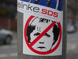 Ein runder Aufkleber auf einer Straßenlaterne mit dem bösen Gesicht von Angela Merkel mit Verbotsymbolik.