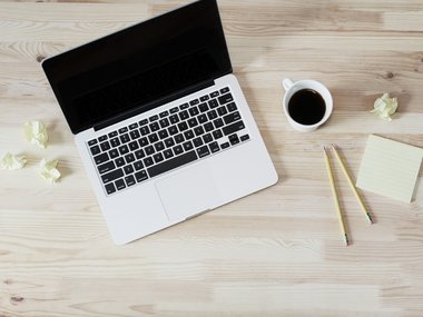 Ein Arbeitsplatz mit Laptop, einer Kaffeetasse, zwei Stiften und zerknüllten Zetteln.