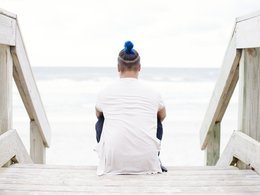 Ein Mann mit blau gefärbtem Haardutt sitzt auf einem Steg zum Strand und blickt in die Ferne.