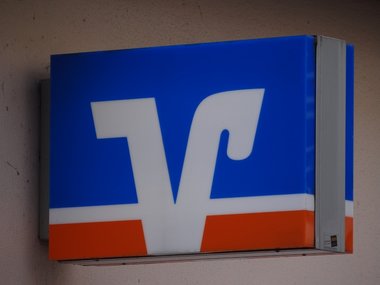 Das blau, weiß und orangene Schild von der Volksbank hängt an einer grauen Wand.