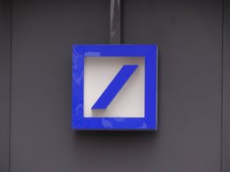 Das blau-weiße Schild von der Deutschen Bank hängt an einer grauen Wand.