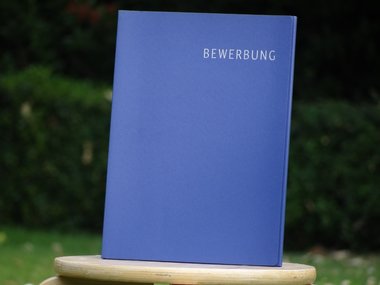 Eine blaue Mappe mit der weißen Aufschrift Bewerbung rechts oben in der Ecke, auf einem Hocker im Garten.