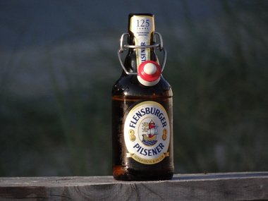Eine Flasche Flensburger Bier steht auf einem Holzbalken mit einem verschwommenem Hintergrund.