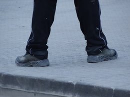 Die breitbeinigen Beine mit Bauarbeiterschuhen auf einem Bürgersteig.