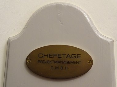 Eine metallfarbiges, ovales Schild von einer Chefetage, Projektmanagement, GMBH.