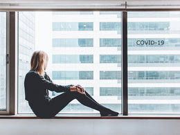 Coronavirus-News: Eine Frau schaut aus einem Bürofenster an dem COVID-19 steht.
