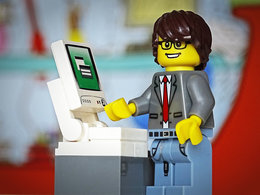 Ein Legomännchen mit Jackett und Krawatte in Jeanshose vor einem Computer symbolisiert ein duales Studium in Wirtschaftsinformatik.