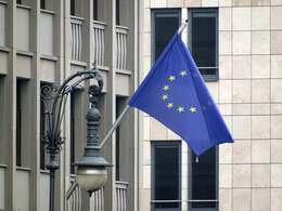 Eine Europaflagge vor einem Betongebäude und eine verschnörkelte Straßenlampe.
