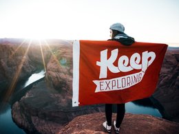 Ein junges Mädchen hält ein rotes Banner mit dem Schriftzug "Keep Exploring" am Grand Canyon hoch.