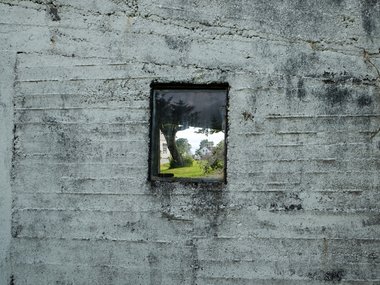 Der Durchblick durch ein Fenster in einer Betonwand auf ein Haus im Grünen.