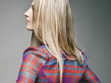 Der Rücken einer Frau mit einer bunt karierten Bluse und langen, blonden Haaren, die zur Seite blickt.