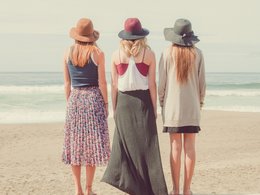 Drei Frauen in weiblicher Kleidung mit Hut stehen mit dem Rücken zum Betrachter gewandt am Strand.