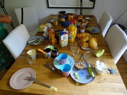 Auf einem Holztisch stehen zahlreiche Lebensmittel und Geschirr für ein Frühstück durcheinander.