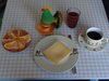 Auf einem kariertem Tisch stehen ein Käsebrötchen, Apfelsine, ein Ei, Saft und Kaffee fürs Frühstück bereit.