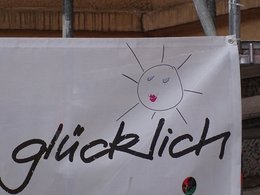 An einem Baugerüst hängt eine weiße Werbeplane. Der fotografierte Teil trägt in Handschrift das Wort glücklich und zeigt eine lachende Sonne. 