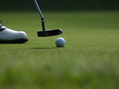 Das Bild zeigt Teile von einem Golfschuh und Golfschläger sowie einen Golfball auf dem grünen Rasen von einem Golfplatz.