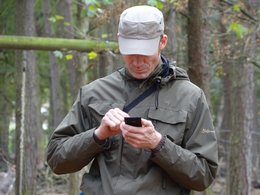 Ein Mann mit Kappi und Jacke steht in einem Wald und tippt auf sein Handy.