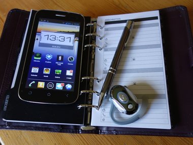 Ein beleuchtetes Smartphone mit zahlreichen Funktionen und Apps und der Uhrzeit: 13.31, sowie einem Notizblock, einem Kugelschreiber und einem Heatset.