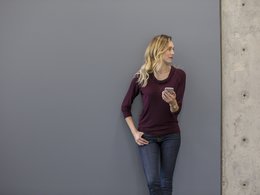 Eine Frau mit blonden, langen Haaren lehnt an einer Wand und hält ein Handy in der Hand.