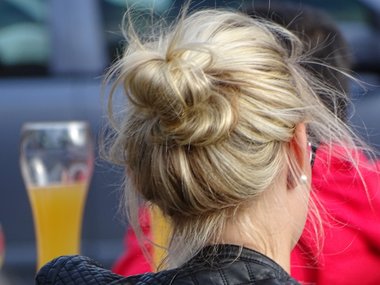 Der Hinterkopf einer Frau mit blonden Haaren, die zu einem Dutt zusammengebunden sind und ein Glas Weizenbier im Hintergrund.