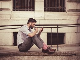 Ein Wirtschaftsstudent sitzt mit angezogenen Beinen, Handy und Kopfhörer auf einer Steinmauer und hört ein Hörbuch.