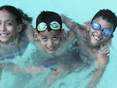 Drei Kinder umarmen sich im Wasser eines Schwimmbades mit und ohne Taucherbrille.