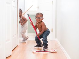 Ein dunkelhäutiges Kind mit freiem Oberkörper und Brille putzt mit einem Wischer den Fußboden.
