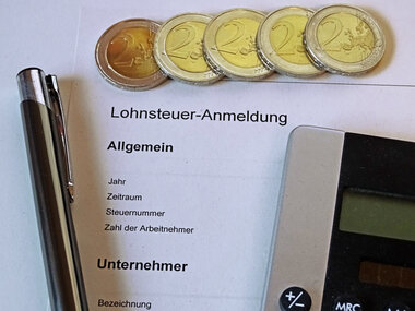 Das Bild zeigt eine Lohnsteuerbescheinigung, einen Taschenrechner, einen Kuli und fünf glänzende 2-Euro Münzen.