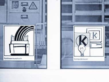 An der Scheibe eines Druckraums zeigen zwei Aufkleber, dass hier Farblaserausdrucker und Computerscanner zur Verfügung stehen.