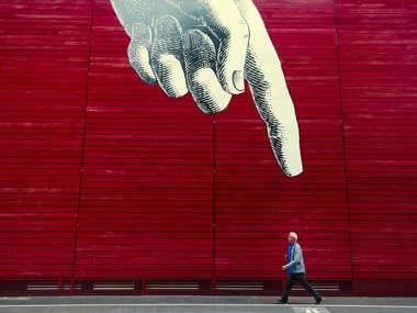 Ein gemalter Zeigefinger an einer roten Wand zeigt auf einen vorbeigehenden Mann.
