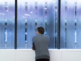 Ein Mann im grauen Pullover stützt sich auf einer Fensterbank ab und schaut nach draußen.