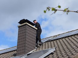 Ein Schornsteinfeger steht auf dem Dach eines Hauses mit Solaranlage am Schornstein.