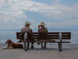 Ein Seniorenpaar mit Strohhut sitzt auf einer Bank am Meer und neben ihnen liegt ein Hund.