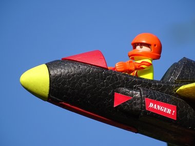 Ein Playmobilmännchen als Flieger mit Helm sitzt in einem Cockpit vor blauem Himmel.