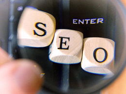 Eine Lupe vergrößert das Wort "SEO" auf drei Holzwürfeln, das für "search engine optimization" also auf Deutsch für Suchmaschinenoptimierung steht.