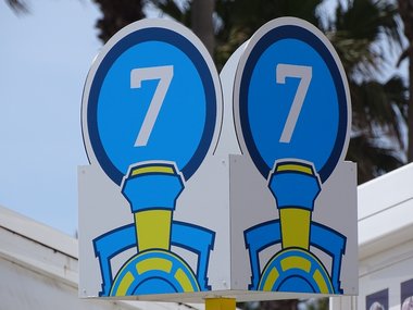 Ein rundes, blaues Schild mit einer 7 in doppelter Ausführung mit einer angedeuteten Eisenbahn darunter.