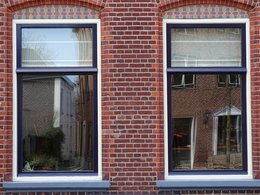 Zwei Fenster einer Wohnungen von einem roten Backsteingebäud mit blauem Rahmen spiegeln den Vorplatz und dessen Häuser wieder.
