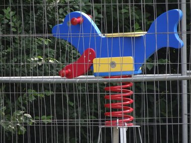 Eine bunte Spielplatzwippe als Ente steht hinter einem Gitterzaun.