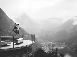 Das schwarz-weiß Bild von einer Aussichtsplattform in ein Tal mit einem Mann, der einen hohen Sprung absolviert.