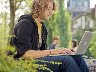 Student sitzt auf dem Campus an der TUM auf der Wiese und arbeitet am Laptop (Mac)