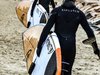 Zwei Surfer in Neoprenanzügen gehen mit dem gleichen Surfbrett unter dem Arm zum Meer.