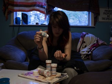 Eine junge Frau sitzt auf einem Sofa in einem düsteren Zimmer und hält Tabletten in der Hand. Mehrere Dosen stehen vor ihr auf dem Tisch.