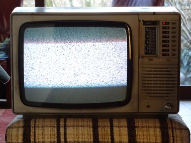 Ein sehr altes TV-Gerät mit Kriselbild, steht auf einem beige-braun gestreiften Hocker.