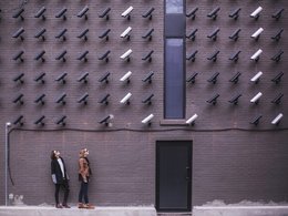 Zahlreiche schwarze und weiße Überwachungskameras sind an einer Gebäudewand angebracht und zwei Frauen mit Sonnenbrille betrachten diese von unten.