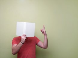 Ein Mann mit rotem Shirt versteckt sein Gesicht hinter einem weißen Buch und hält den Zeigefinger der freien Hand nach oben.