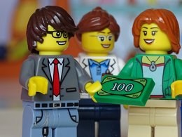 Banker - Das Bild zeigt Lego-Männchen: Ein Banker übergibt Geld an seine Kundinnen.