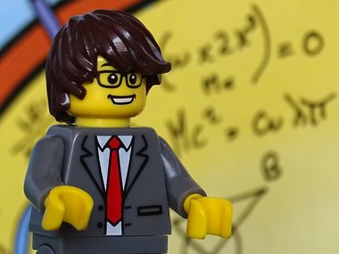 Wirtschaftsmathematiker - Das Bild zeigt ein Lego-Männchen im Anzug vor einer Wand mir lauter Zahlen Pi und geometrischen Formen. 